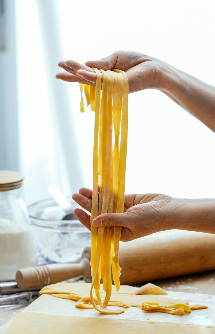 Unrecognizable person with long uncooked pasta standing at table with rolling pin while cooking tagliatelle with dough in kitchen against blurred background