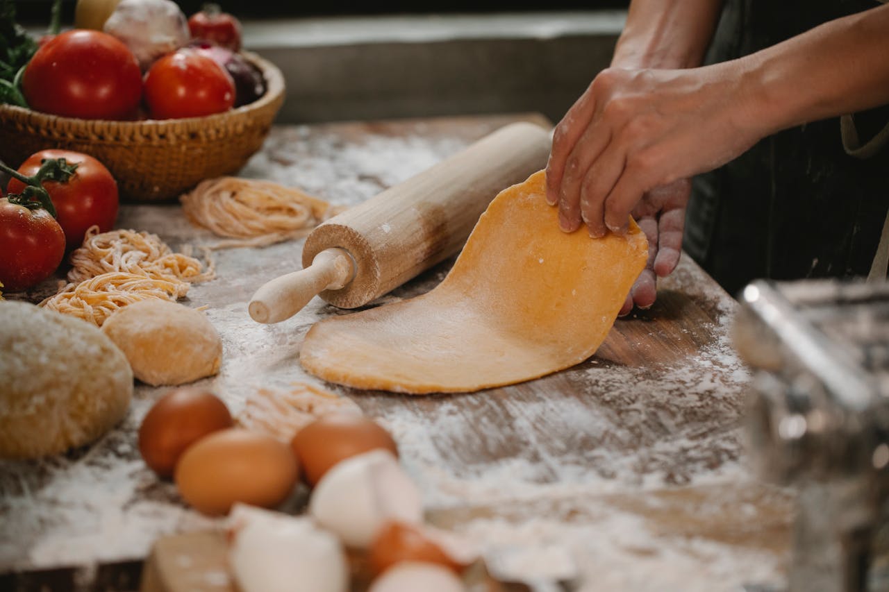 High angle of crop faceless female kneading dough with rolling pin on wooden floury table with scattered eggs and vegetables while preparing pasta